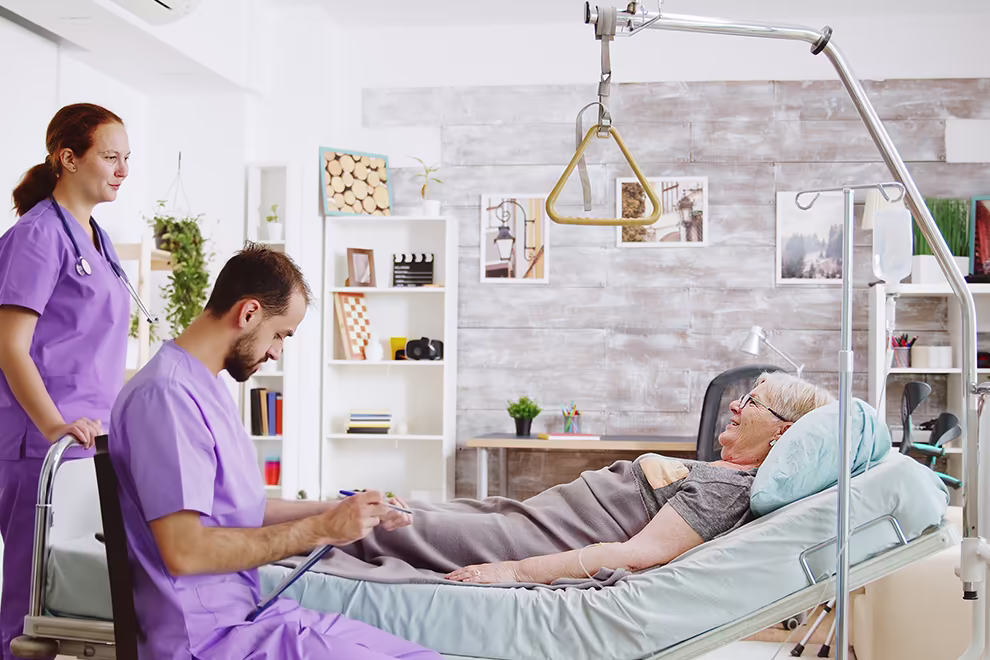 mittendrin-01-990 Zwei Pflegefachkräfte stehen bei einem ambulanten Pflegebesuch an einem Bett