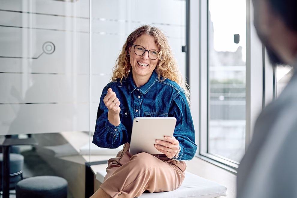 mittendrin-03-990 Eine Frau sitzt in einem Büro auf einer Fensterbank aus Holz und hält ein Tablet in der Hand